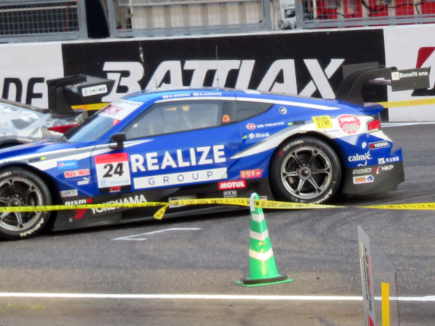 Ein blau-weißer Rennwagen fährt auf einer Rennstrecke, mit einem Verkehrskegel im Vordergrund und Tafeln mit Text, Geländern und anderen Gegenständen im Hintergrund.