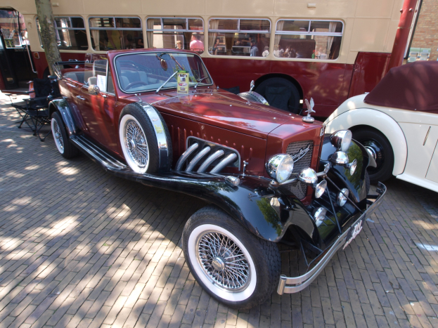 Vintage-Auto neben einem Doppeldeckerbus auf einem Kopfsteinpflasterweg geparkt, mit Stühlen und einem Baum im Vordergrund, einem Gebäude im Hintergrund und Menschen im Bus sichtbar.