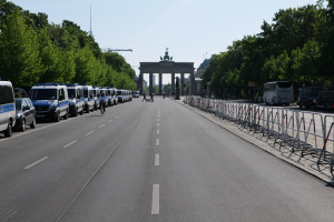 Eine Reihe von Polizeiwagen auf einer Straße vor dem Brandenburger Tor in Berlin geparkt, mit Menschen auf Fahrrädern und zu Fuß, Barrieren, Bäumen und einem Bogen mit Statuen im Hintergrund.