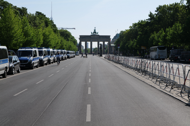 Eine Reihe von Polizeiwagen auf einer Straße vor dem Brandenburger Tor in Berlin geparkt, mit Menschen auf Fahrrädern und zu Fuß, Barrieren, Bäumen und einem Bogen mit Statuen im Hintergrund.