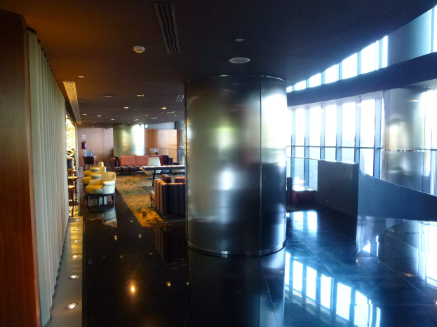 Eine geräumige First-Class-Lounge am Hong Kong International Airport mit großen Fenstern, hohen Decken mit elektrischem Licht, Sitzmöglichkeiten einschließlich Stühlen und Sofas, Tischen, einem Teppichboden und einer mittigen Säule.