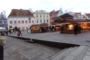 Ein lebendiger Weihnachtsmarkt in Tallinn, Estland, mit Menschen um beleuchtete und geschmückte Stände, Gebäuden mit Fenstern im Hintergrund, Treppen, die zum Markt führen, und Töpfen mit Pflanzen drumherum.