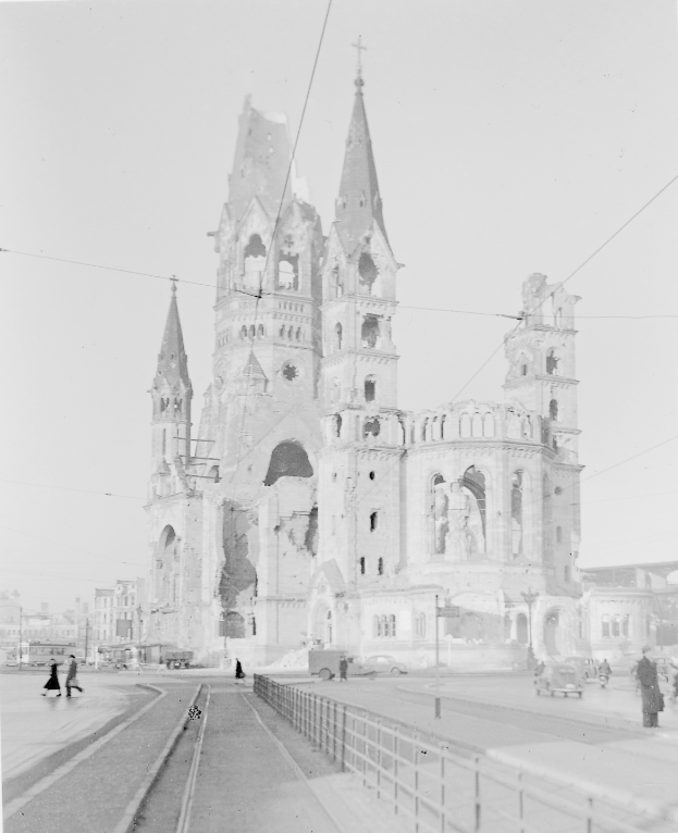 Ein Schwarz-Weiß-Foto einer Kirche in einer Stadt, umgeben von Gebäuden, Straßenlaternen, Straßenlaternen, Kraftfahrzeugen, Fußgängern, Geländern, Strommasten, Stromkabeln und einem klaren Himmel.