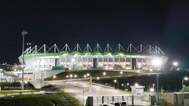 Ein großes Stadion bei Nacht beleuchtet mit parkenden Autos davor, umgeben von Straßenmasten, Straßenlaternen, Strommasten, Stromkabeln, Kraftfahrzeugen auf der Straße, Gittern, Bäumen und einem sternenklaren Himmel.