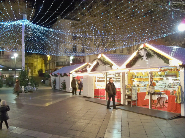 Ein lebendiger Weihnachtsmarkt Nachts In Einer Stadt, Mit Menschen, Die Stehen, Sitzen Und Ein Kind Halten, Umgeben Von Festlichen Lichtern, Pfählen, Pflanzen, Bäumen Und Gebäuden Mit Fenstern Unter Einem Wolkenverhangenen Himmel.
