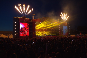 Eine große Menschenmenge versammelte sich auf einem Musikfestival, mit einer Bühne in der Mitte, auf der Musiker im Scheinwerferlicht spielten und von Bannern umgeben waren, Zelte links und ein dunkler Himmel mit Feuerwerk im Hintergrund.