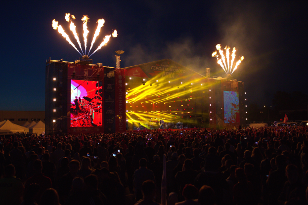 Eine große Menschenmenge versammelte sich auf einem Musikfestival, mit einer Bühne in der Mitte, auf der Musiker im Scheinwerferlicht spielten und von Bannern umgeben waren, Zelte links und ein dunkler Himmel mit Feuerwerk im Hintergrund.