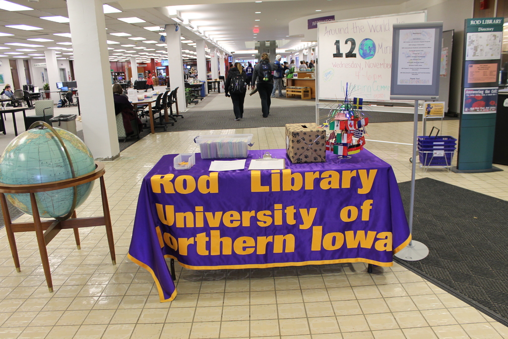Eine Ausstellung mit einem Tisch, auf dem verschiedene Landesflaggen, ein Karton, Papiere und ein Banner liegen, eine Kugel zur Linken, ein Anschlagbrett zur Rechten mit einigen Menschen in der Nähe und mehrere Tische und Stühle ganz links.