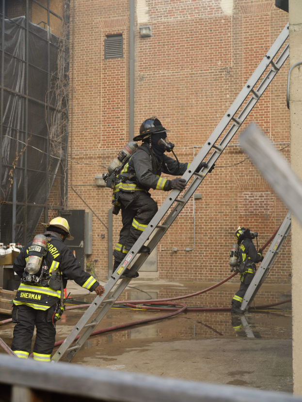 Feuerwehrleute in Helmen und Lufttanks klettern an einer Leiter vor einem Backsteingebäude mit Rohren auf dem Boden und einem Metallstab am unteren Ende, mit einem weiteren Gebäude mit Fenstern und Netz im Hintergrund.