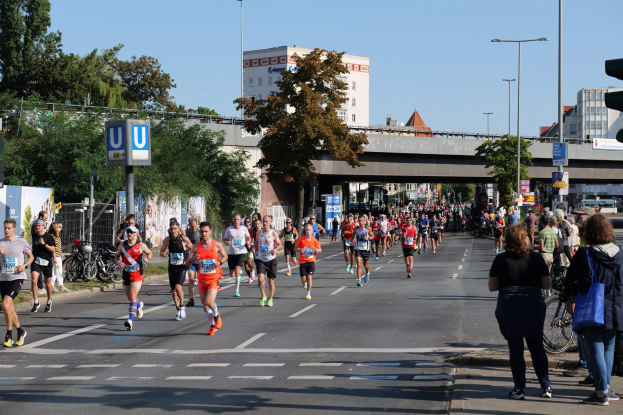 Gruppe von Menschen, die einen Marathon auf einer von Bäumen gesäumten Straße mit Fahrrädern, Schildern, einem Zaun, Gras, einer Brücke, Gebäuden und einem klaren blauen Himmel laufen.