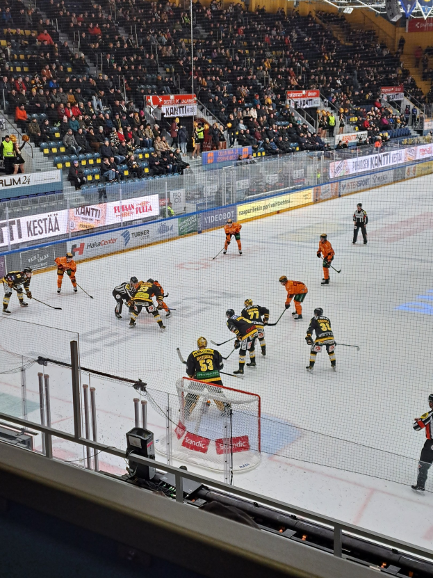 Eine Gruppe von Menschen spielt Hockey auf einem Eisstadion, umgeben von einem Zaun, mit Zuschauern im Hintergrund und beleuchtet von Scheinwerfern.