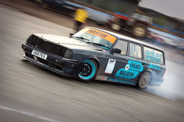 Ein Auto rast auf einer Rennstrecke, wobei Rauch aufsteigt, umgeben von anderen Fahrzeugen und Zuschauern.