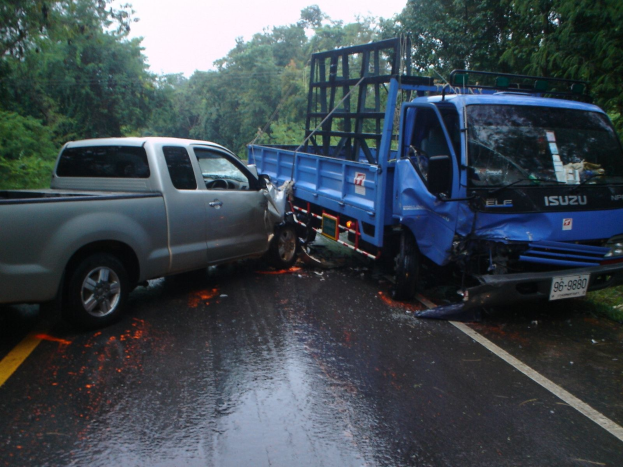 Ein schwerbeschädigter Lkw mit eingedrückter Front und eingebeulter Karosserie liegt auf der Seite einer Straße, umgeben von Bäumen unter einem klaren blauen Himmel.