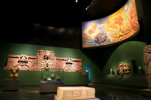Eine Museumsausstellung im Anthropologischen Museum von Mexiko-Stadt, die verschiedene Kunstwerke zeigt, darunter Skulpturen, Objekte auf Tischen, eine Texttafel, eine Wandmalerei und Deckenleuchten.