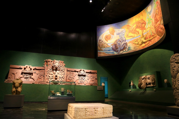 Eine Museumsausstellung im Anthropologischen Museum von Mexiko-Stadt, die verschiedene Kunstwerke zeigt, darunter Skulpturen, Objekte auf Tischen, eine Texttafel, eine Wandmalerei und Deckenleuchten.