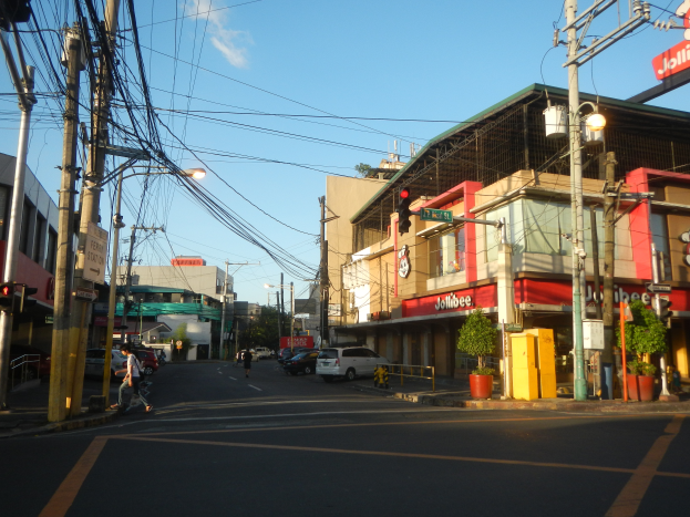 Eine Stadtstraße mit Autos, Fußgängern, Strommasten, Laternen, Ampeln, Gebäuden mit Namensschildern, Topfpflanzen und einer bewölkten Himmel.