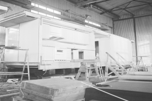 Ein Schwarz-Weiß-Foto eines Gebäudes im Bau mit Leitern, Holzboxen und anderen Gegenständen, die auf dem Boden verstreut sind, und einer Wand mit Fenstern und Lichtern im Hintergrund.