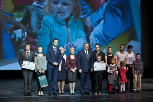 Gruppe von Menschen auf einer Bühne mit Taschen und Urkunden in den Händen, im Hintergrund ein Bildschirm mit einem Kind und anderen.