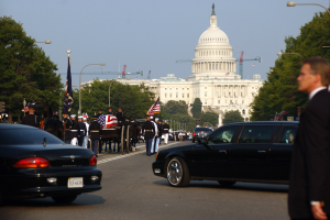 Eine Trauerprozession mit Menschen in Mützen und mit Fahnen zieht am Kapitolsgebäude vorbei, flankiert von Laternenmasten, Bäumen und Gebäuden unter einem klaren Himmel.