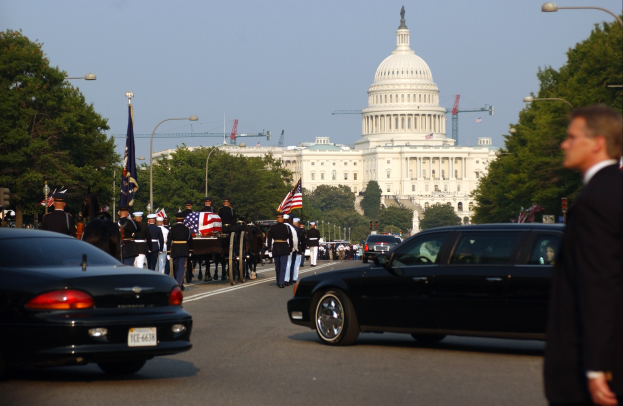 Eine Trauerprozession mit Menschen in Mützen und mit Fahnen zieht am Kapitolsgebäude vorbei, flankiert von Laternenmasten, Bäumen und Gebäuden unter einem klaren Himmel.