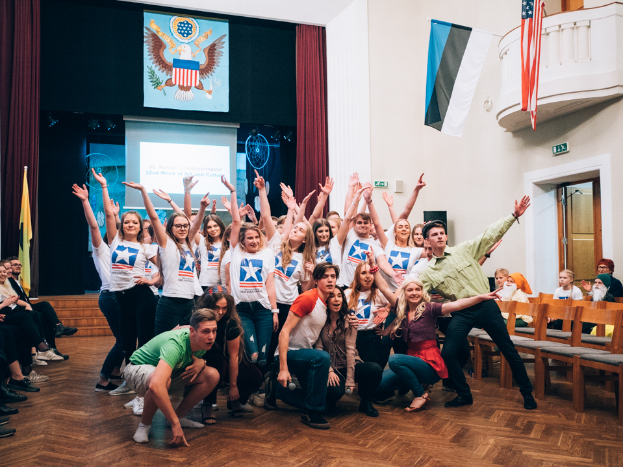 Eine Gruppe von Menschen posiert vor einer Menge, die auf Stühlen sitzt und steht, mit einem Bildschirm, Vorhängen, Flaggen, einem Schild, einer Tür, einem Geländer und einer Decke mit Deckenleuchten im Hintergrund.