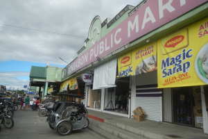 Eine belebte Stadtstraße mit parkenden Fahrzeugen, Fußgängern, Gebäuden, Strommasten, Bäumen und einem bewölkten Himmel, mit einem Geschäft mit der Aufschrift "Bongabon Public Market" im Vordergrund.
