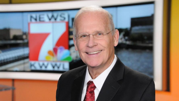 Ein Mann in Anzug und Krawatte, mit Brille, steht lächelnd vor einem Fernseher, mit einem unscharfen Hintergrund.