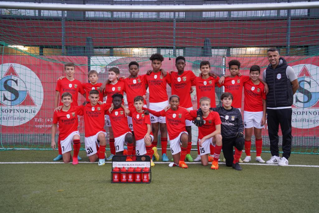 Eine Gruppe junger Männer in roten T-Shirts und schwarzen Shorts steht auf einem Fußballfeld vor einer Kiste roter Gegenstände, im Hintergrund Schilder mit der Aufschrift "FC Rot Schwarz Eglaute - Champions League"