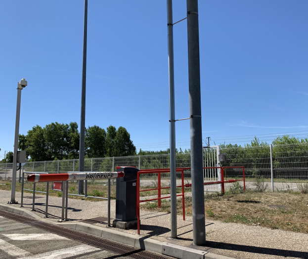 Ein rotes und weißes Schild an einem Bahnhof, umgeben von einem Metallzaun, Pfählen, Bäumen, Pflanzen, Gras, einem Straßenpfahl, Drähten und einem bewölkten Himmel, mit einer Absperrung im Vordergrund.