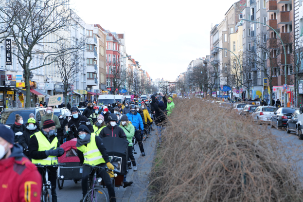 Eine große Gruppe von Menschen in Masken und Sicherheitswesten auf Fahrrädern eine von Bäumen gesäumte Straße mit Gebäuden, Fahrzeugen, Laternen und Texttafeln entlang, unter einem klaren blauen Himmel.
