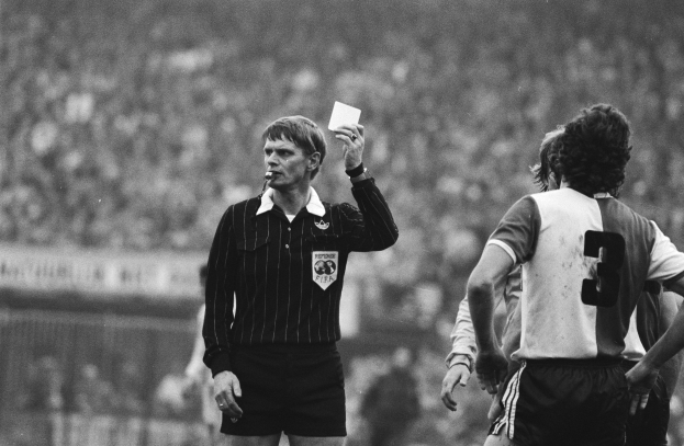 Schwarzes und weißes Bild eines Schiedsrichters mit roter Karte vor einer Menge von Menschen in Fußballuniformen, mit einem Zaun im Hintergrund.