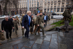 Boris Johnson beim Eintreffen im Kreml in Moskau, vorbei an einer Gruppe von Menschen auf einem Gehweg neben einem Gebäude, einige tragen Helme und halten Waffen, Fahrzeuge auf der Straße und Gebäude im Hintergrund.