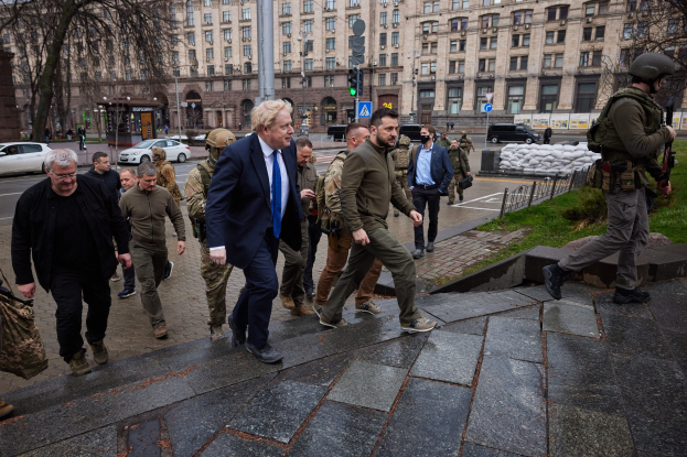 Boris Johnson beim Eintreffen im Kreml in Moskau, vorbei an einer Gruppe von Menschen auf einem Gehweg neben einem Gebäude, einige tragen Helme und halten Waffen, Fahrzeuge auf der Straße und Gebäude im Hintergrund.
