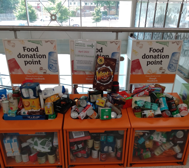 Orange Schränke mit gespendeten Lebensmitteln wie Flaschen, Dosen und Kartons, mit Informationsschildern in der Nähe, durch Glasfenster betrachtet, die Fahrzeuge, Bäume, Gebäude und Himmel zeigen.