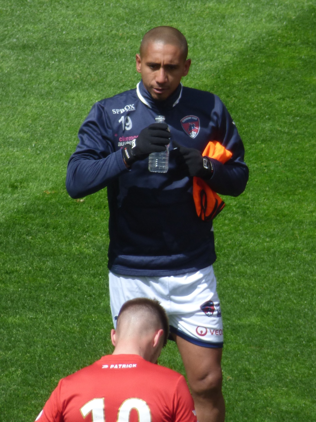 Ein professioneller Athlet steht auf einem saftig grünen Feld, hält in einer Hand eine Flasche und in der anderen ein oranges Tuch.