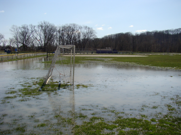 Ein Fußballtor steht in der Mitte eines überfluteten Feldes, mit Gras, einem Zaun, Bäumen, Häusern und einem bewölkten Himmel im Hintergrund.