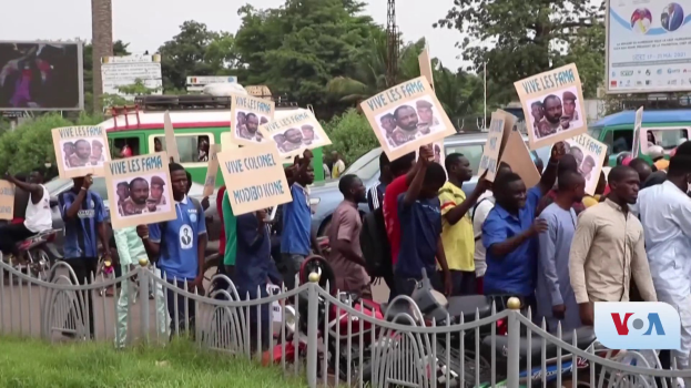 Eine Gruppe von Menschen h├Ąlt Schilder in H├Ąnden und steht vor einem Zaun, umgeben von Fahrzeugen, B├Ąumen, Masten und Plakaten, mit einem Bildschirm und Gras im Hintergrund, wahrscheinlich protestieren sie gegen eine Regierungsentscheidung, die Video-Konferenzen verbietet.