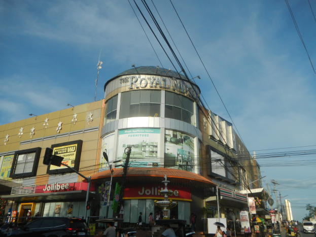 Eine belebte Stadtstraße mit fahrenden Autos, Fußgängern auf dem Gehweg und dem Royal Mall Einkaufszentrum im Hintergrund, umgeben von Bäumen, Strommasten und Drähten unter einem Himmel.