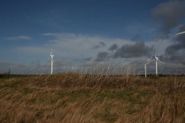 Luftaufnahme eines Windparks mit mehreren Turbinen auf einer grünen Wiese, umgeben von Bäumen und Wolken, mit "Niederlande" unten sichtbar.