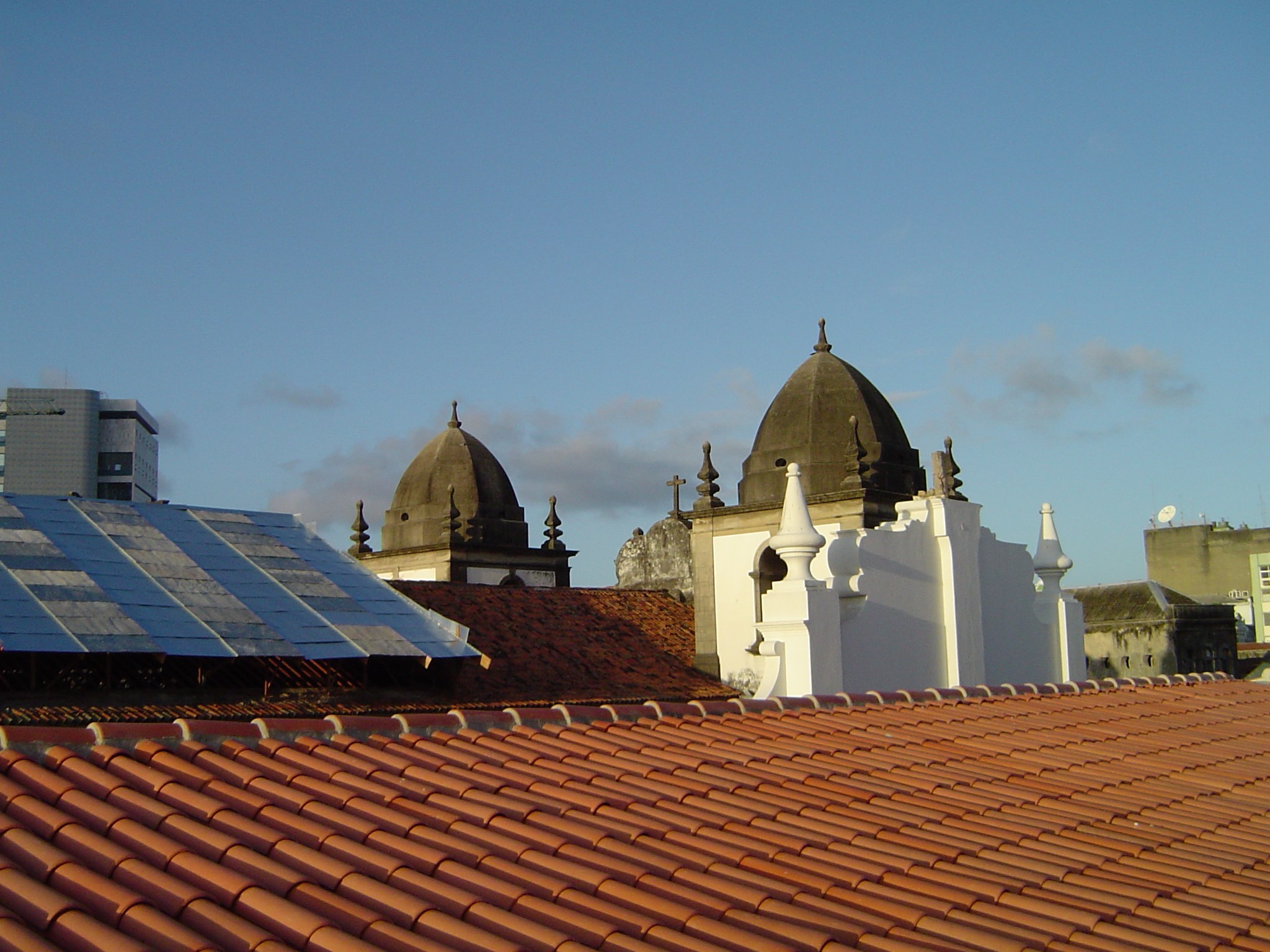 Stadtpanorama mit Gebäuden im Vordergrund und einem blauen Himmel im Hintergrund, mit Solarpanelen auf einem Dach, die den Einsatz erneuerbarer Energien anzeigen.