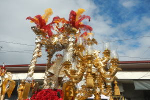 Ein großer goldener und roter Festwagen, der mit Blumen und anderen Schmuckelementen geschmückt ist, in einem Karnevalsumzug mit einem Gebäude, Strommasten mit Drähten und einem bewölkten Himmel im Hintergrund.