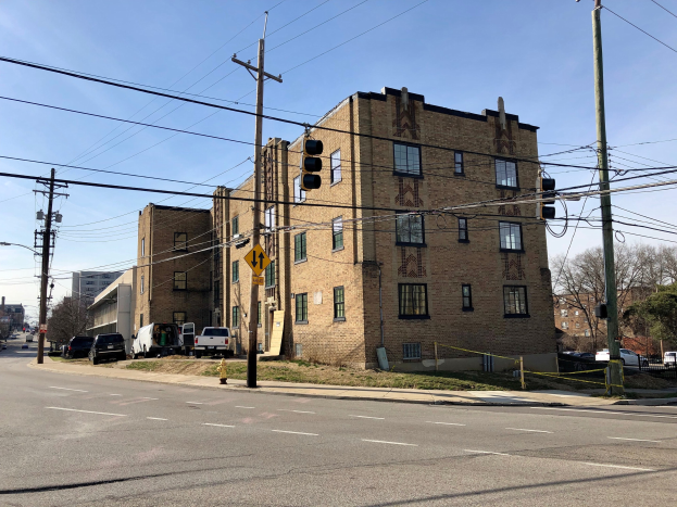 Großer Ziegel-Eckbau mit mehreren Fenstern, umgeben von städtischen Elementen einschließlich Strommasten, Ampeln, Fahrzeugen, Bäumen und einem bewölkten Himmel.