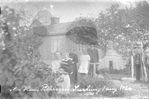 Ein altes Schwarz-Weiß-Foto einer Familie, die vor einem Haus steht, mit einer Frau, die ein Baby hält, anderen Familienmitgliedern drumherum, Bäumen und einer Hütte im Hintergrund und Text unten.