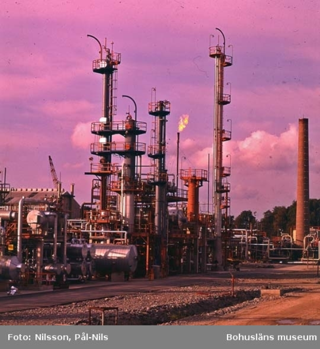 Ein Ölraffinerie mit zahlreichen Rohren, Ventilen, Metallkonstruktionen, Containern, einem Gebäude, einem Kran und einer Gruppe von Bäumen unter einem bewölkten Himmel, mit Text am unteren Bildrand.