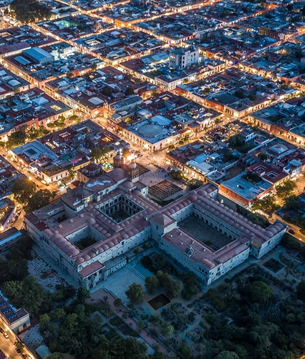 Luftaufnahme einer nächtlichen Stadt mit beleuchteten Gebäuden, Bäumen, Straßen und Fahrzeugen, die ein großes zentrales Gebäude zeigt, das wie ein Gefängnis aussieht.