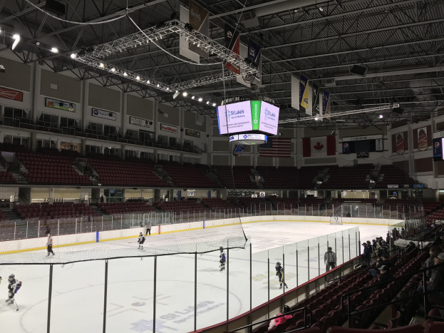 Eishockeyspiel in einer großen Arena mit einem zentralen Tor, Zuschauern auf Stühlen, Decke mit Lampen und Trägern sowie Bannern an den Wänden, identifiziert als die St. Louis Blues Arena.
