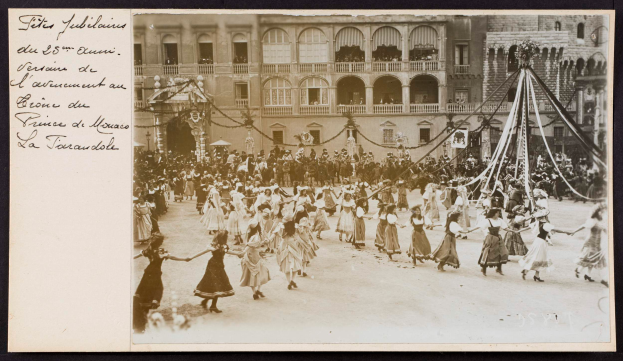 Ein Schwarz-Weiß-Foto von Menschen in traditioneller Tracht, die vor einem Gebäude mit Fenstern, Bögen, Säulen und Geländern tanzen, mit einem Pfahl mit Gegenständen rechts und Text links.