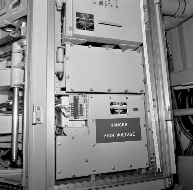 Ein Schwarz-Wei├č-Foto eines elektrischen Schaltpults mit einer Gefahrenwarnung, umgeben von verschiedenen Maschinen und Rohren.