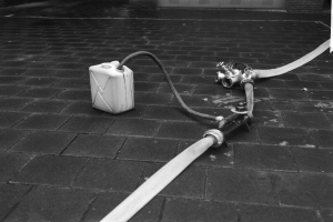 Schwarz-weiß-Foto eines Gasbehälters mit einem Schlauch, der vor einem Gebäude mit einer Tür im Hintergrund auf dem Boden steht.