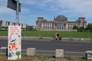 Eine Person in einem schwarzen Kleid f√§hrt mit dem Fahrrad die Stra√e vor dem Reichstag in Berlin, Deutschland, entlang, mit Geb√§uden, B√§umen und Menschen im Hintergrund.